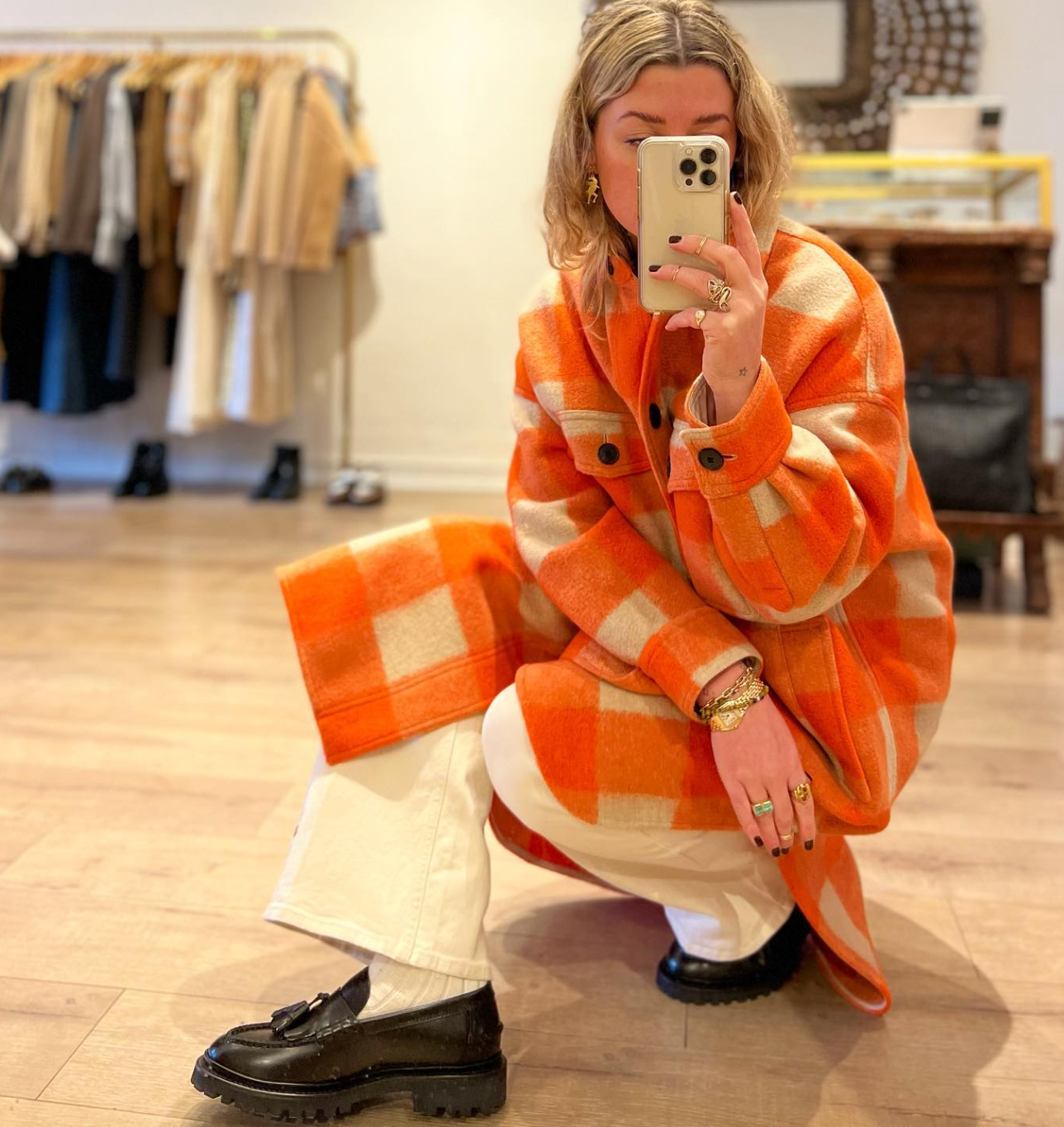 Eli squatting in the store wearing an orange checked jacket with white jeans and loafer